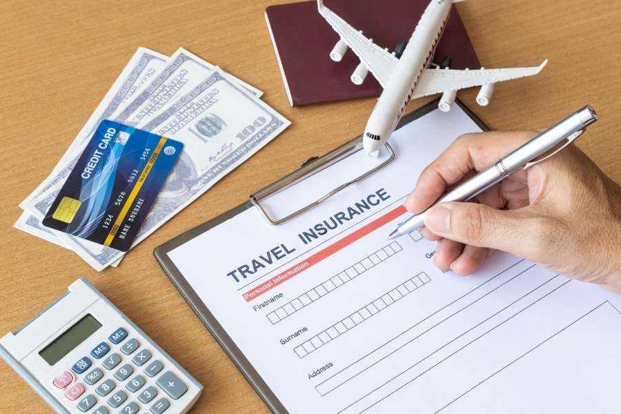 Une main remplit un formulaire d'assurance voyage sur un bureau avec un modèle d'avion, de l'argent, une carte de crédit et un passeport.