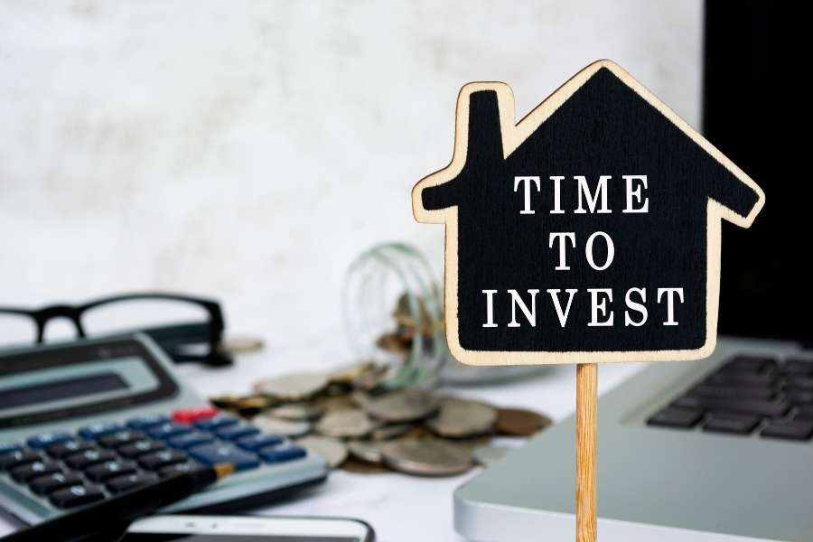 Un panneau en forme de maison avec l'inscription "Time to invest" posé à côté d'une calculatrice et de pièces de monnaie pour illustrer comment investir en colocation.